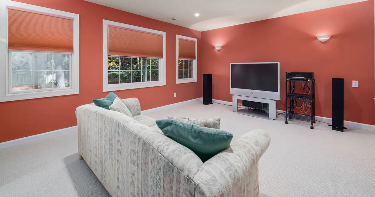 Spacious home theater room with orange walls and a comfortable couch, featuring a flat-screen TV and ample natural light