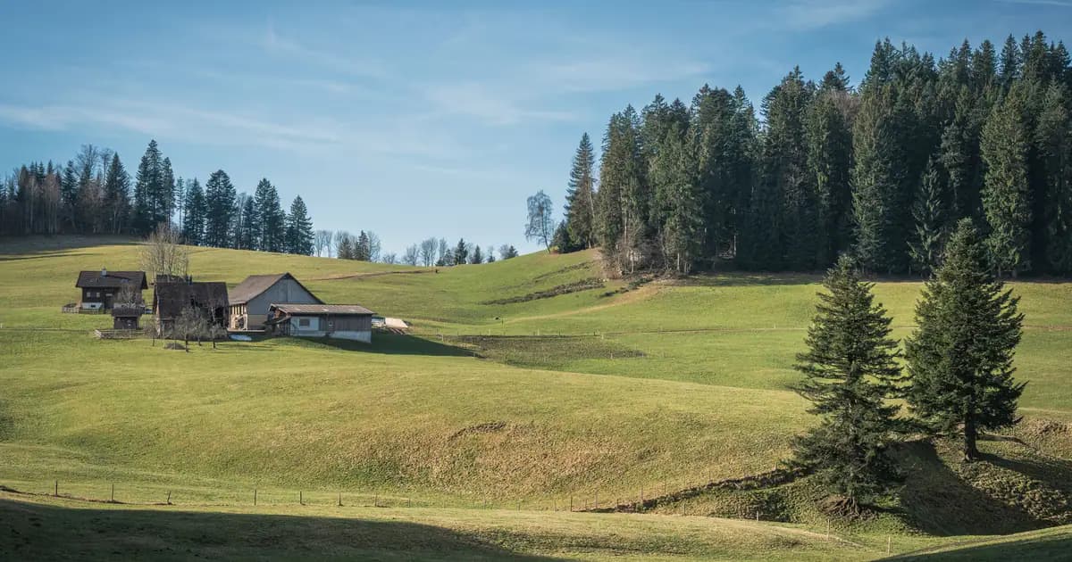 Picturesque rural landscape featuring farmhouses, rolling hills, and dense trees under a clear blue sky