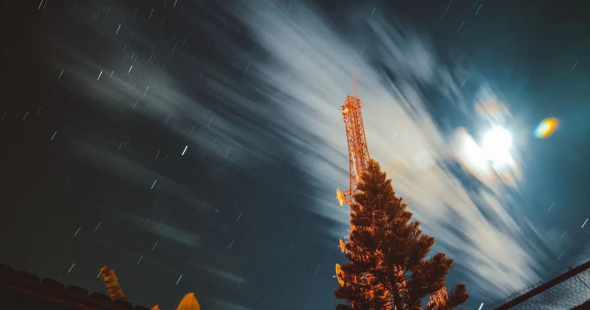 Long exposure of a night sky featuring an antenna with star trails and clouds