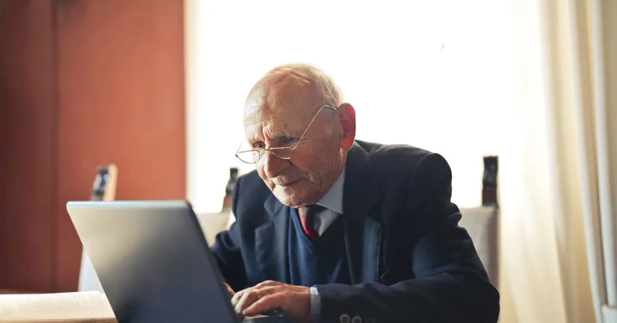Concentrate elderly senior man in formal suit and eyeglasses working on laptop while sitting at wooden table with books