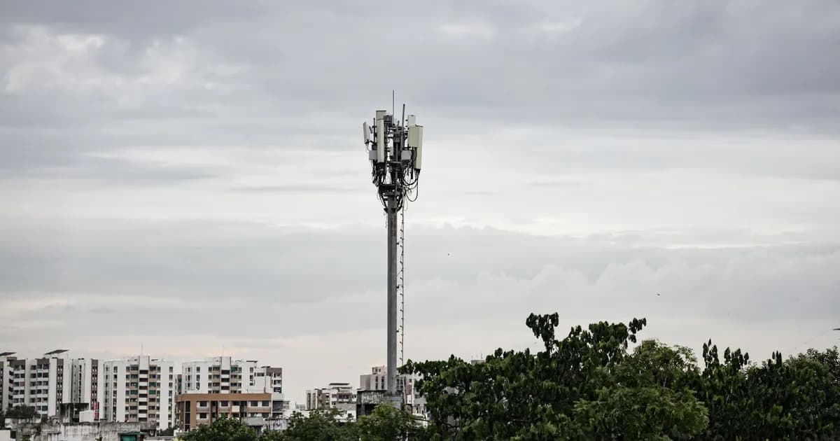 Cell tower rising above trees with urban high-rise buildings under a cloudy sky