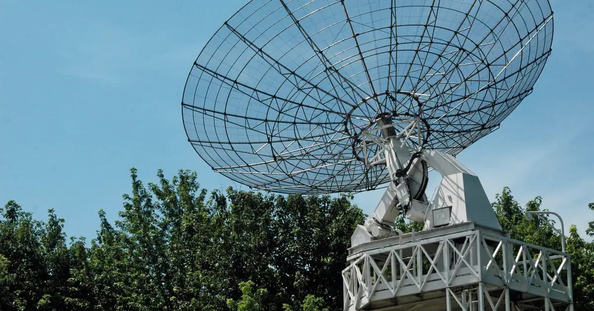A large satellite dish stands tall against a clear blue sky, surrounded by greenery