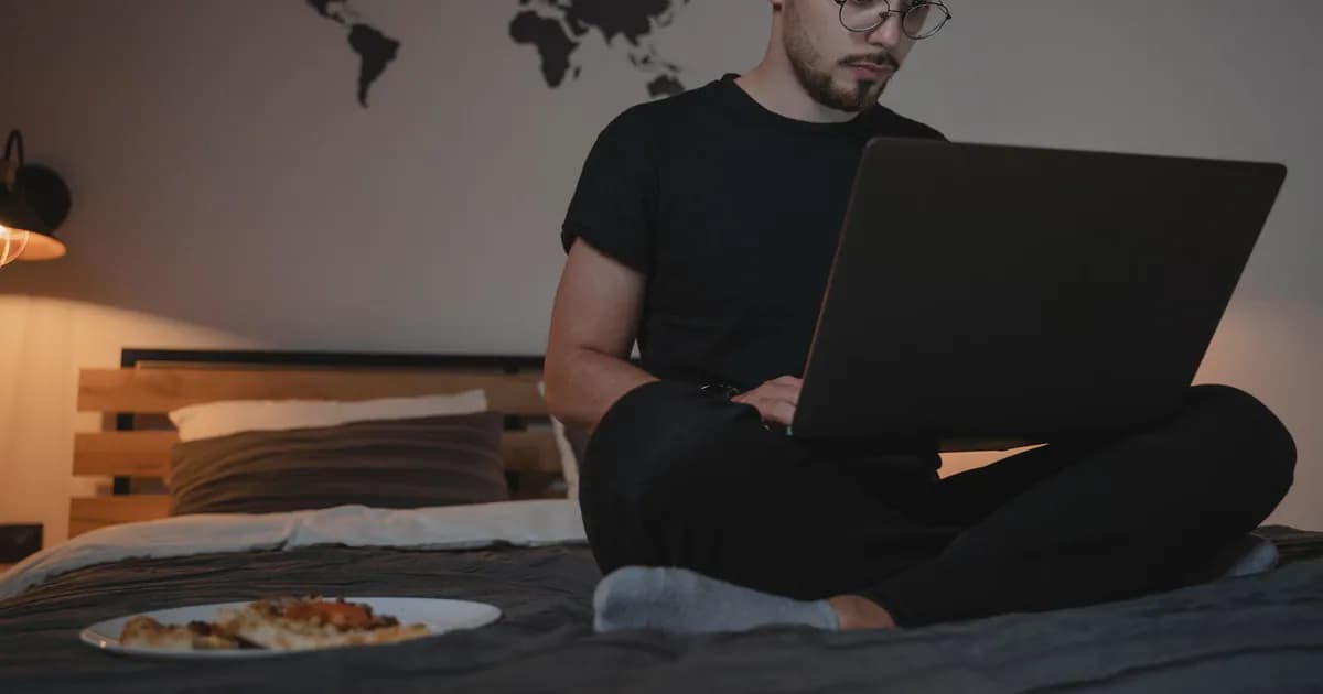 A man sits on a bed with a laptop, working remotely, with a world map and pizza plate nearby