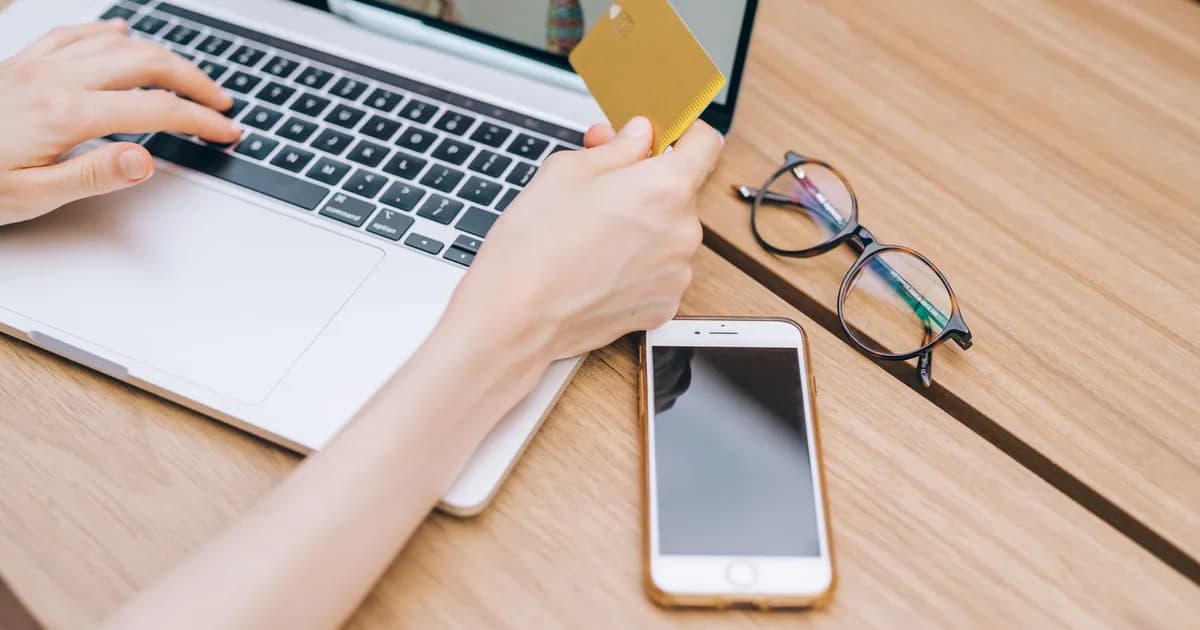 A person makes an online purchase using a credit card on a laptop, with a smartphone and glasses nearby