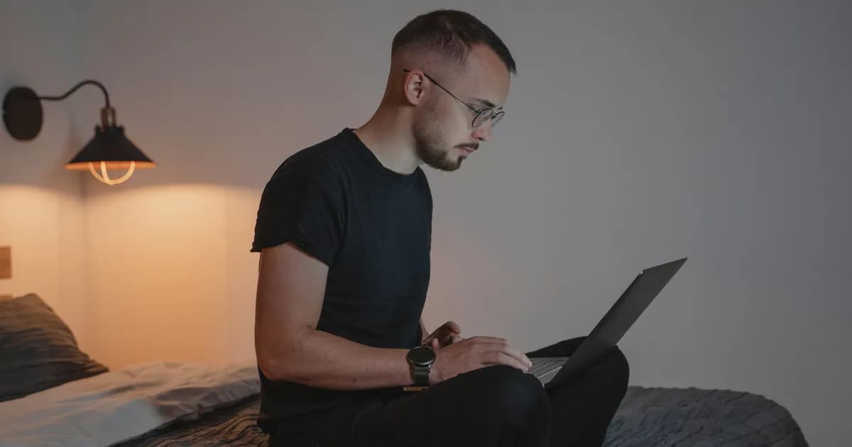 A man in a black shirt sits on a bed using a laptop, with a plate of food nearby