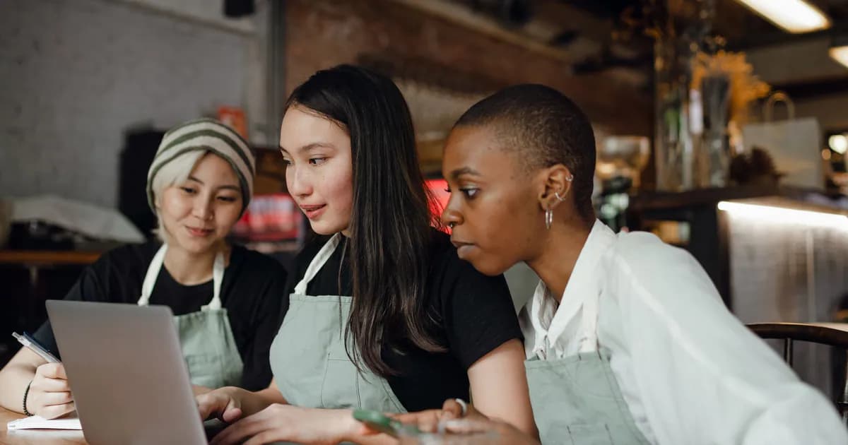 Optimistic multiethnic female coworkers in casual clothes and aprons sitting at wooden table in contemporary cafe