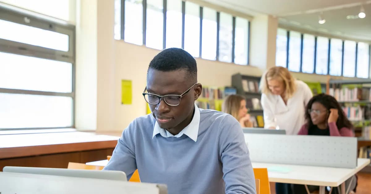 Concentrated young black male student in casual clothes and eyeglasses using laptop sitting at desk in classroom