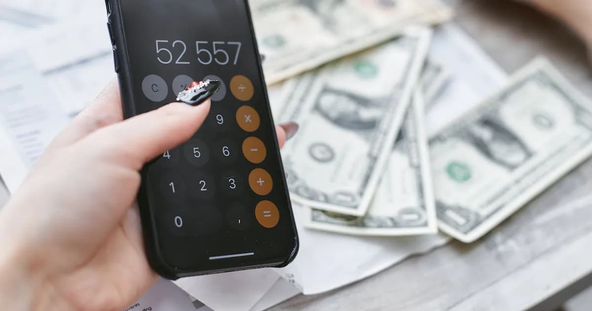 Close-up of hands using smartphone calculator next to dollar bills on table. Financial planning concept