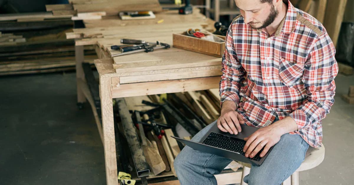 A carpenter working on his laptop in a wood workshop, surrounded by tools and materials