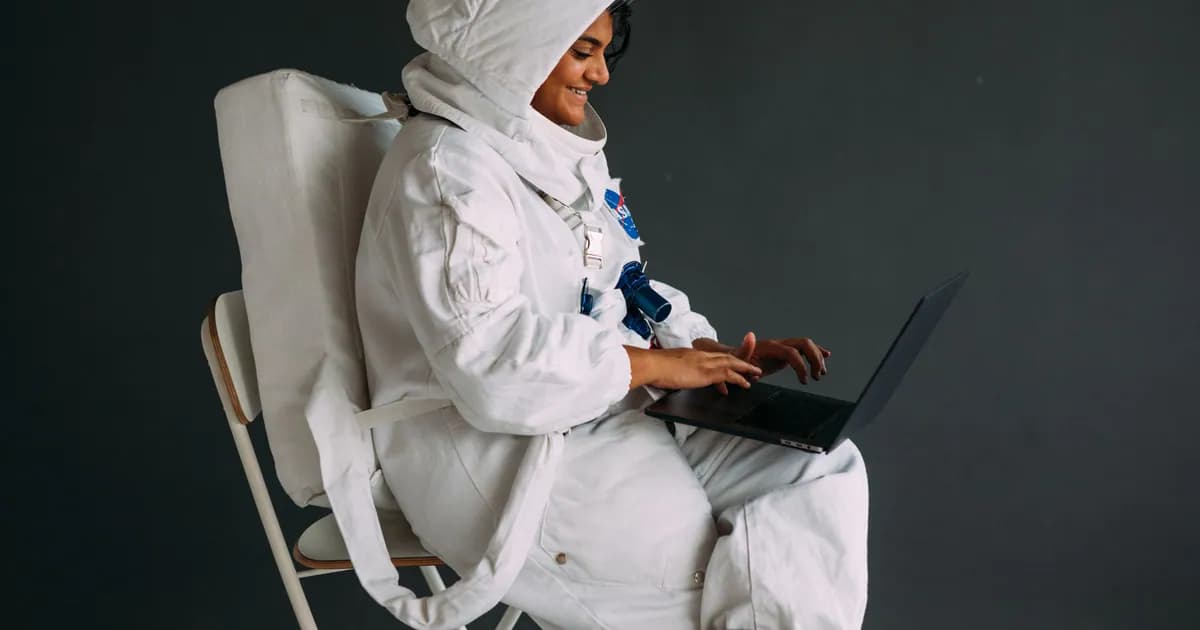 Smiling astronaut in white space suit sits indoors using a laptop