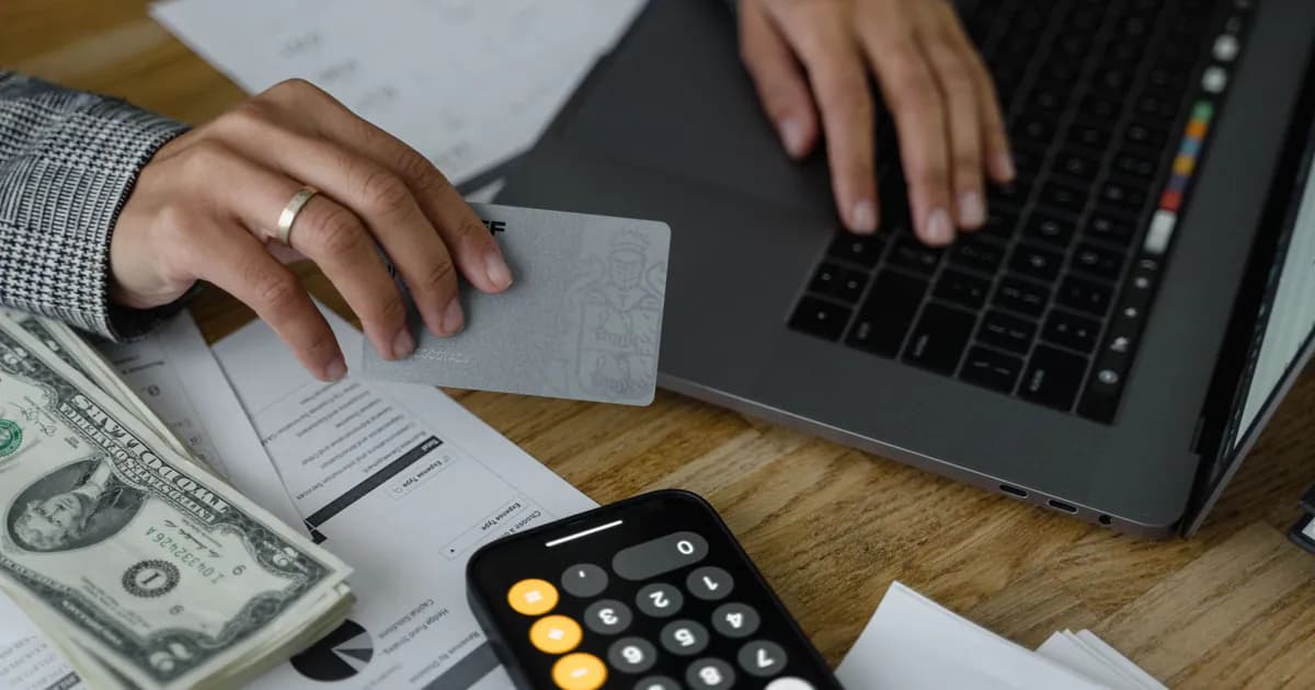 Close-up of a person using a credit card and laptop in a financial setting with cash and calculator