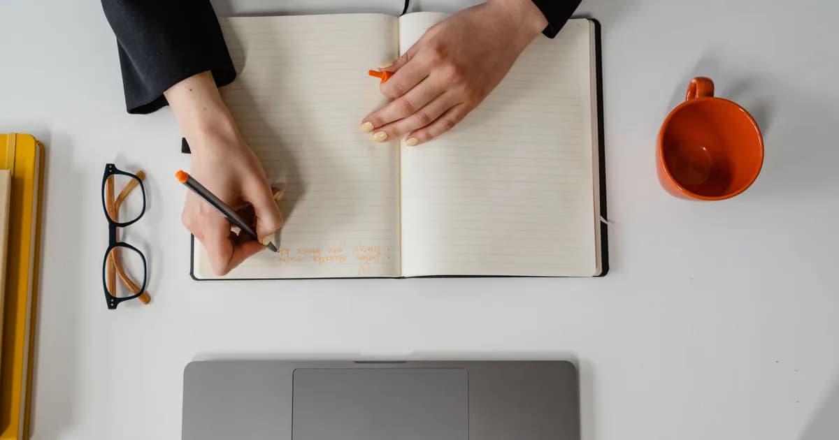 A businesswoman writes notes in a notebook with a laptop, glasses, and mug in view