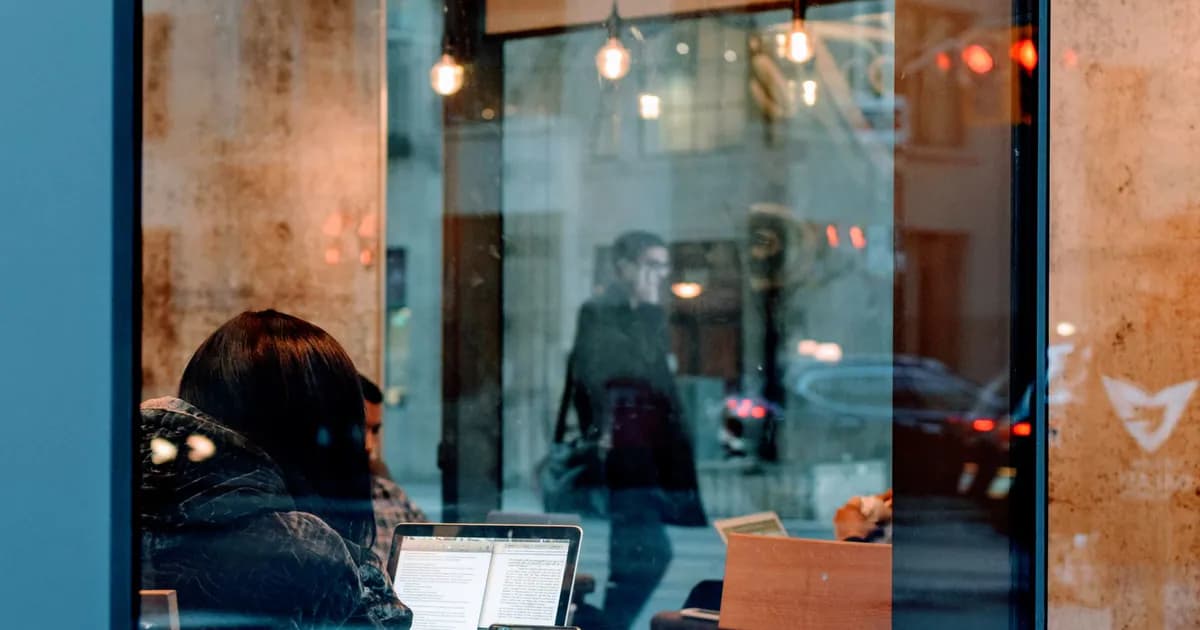 A person working on a laptop inside a café, viewed through a window with street reflections